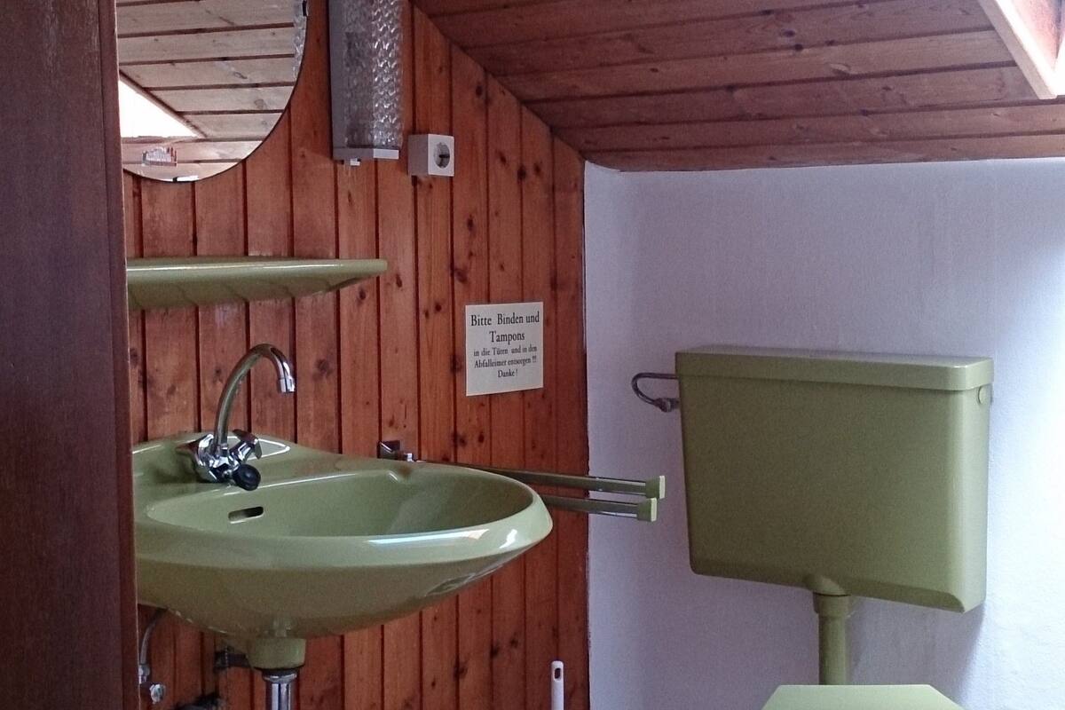 Ein kleines Badezimmer mit Holzboden, einem grünen Waschbecken, einer Toilette und einem kleinen Fenster.