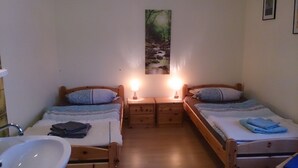 Een kleine, gezellige slaapkamer met twee enkele bedden, een nachtkastje ertussen, een wandplank en een badkamer met een wastafel en spiegel.