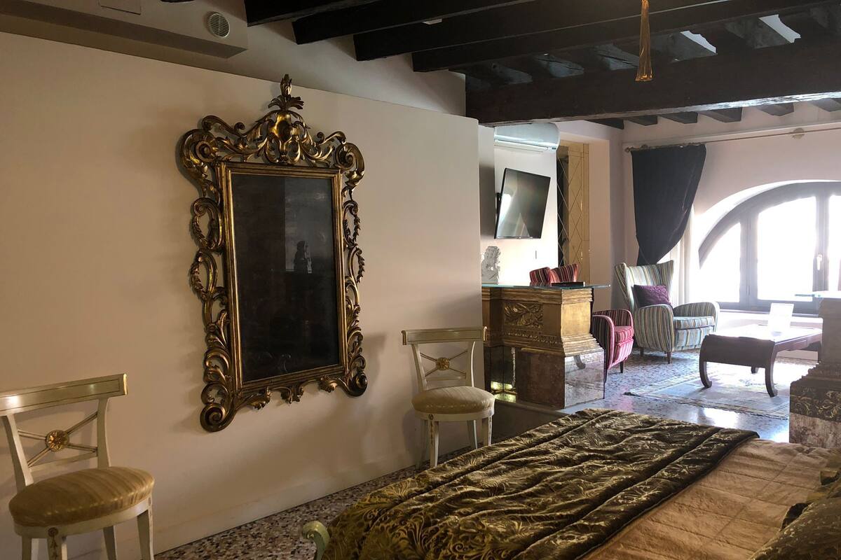 A room with a large ornate mirror, a bed with a patterned bedspread, two upholstered chairs, a small wooden cabinet, and a television on a stand.