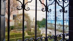 A view through ornate iron gates of a garden with a statue, a building with blue accents, and a stone fountain. - Granda Sweet Suites (Venice)