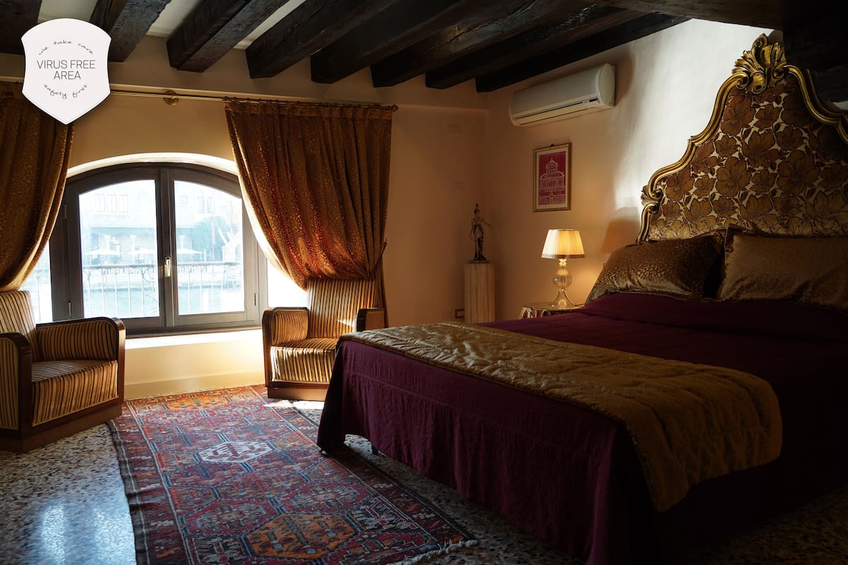 A bedroom with a large bed, a window with curtains, a nightstand with a lamp, and a decorative headboard.