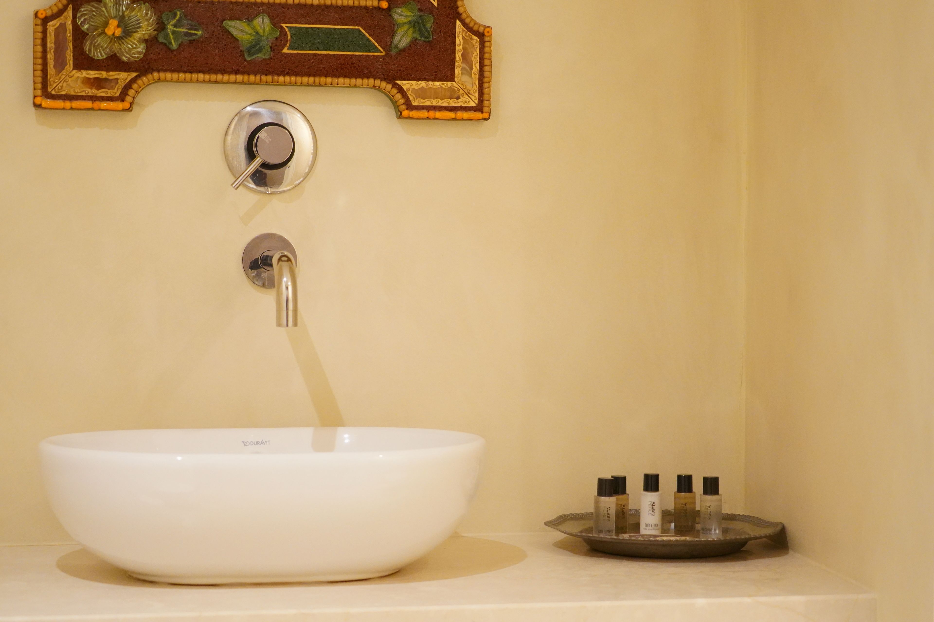 A white vessel sink with a single faucet, a decorative wall plaque above, and a tray with several bottles to the right.