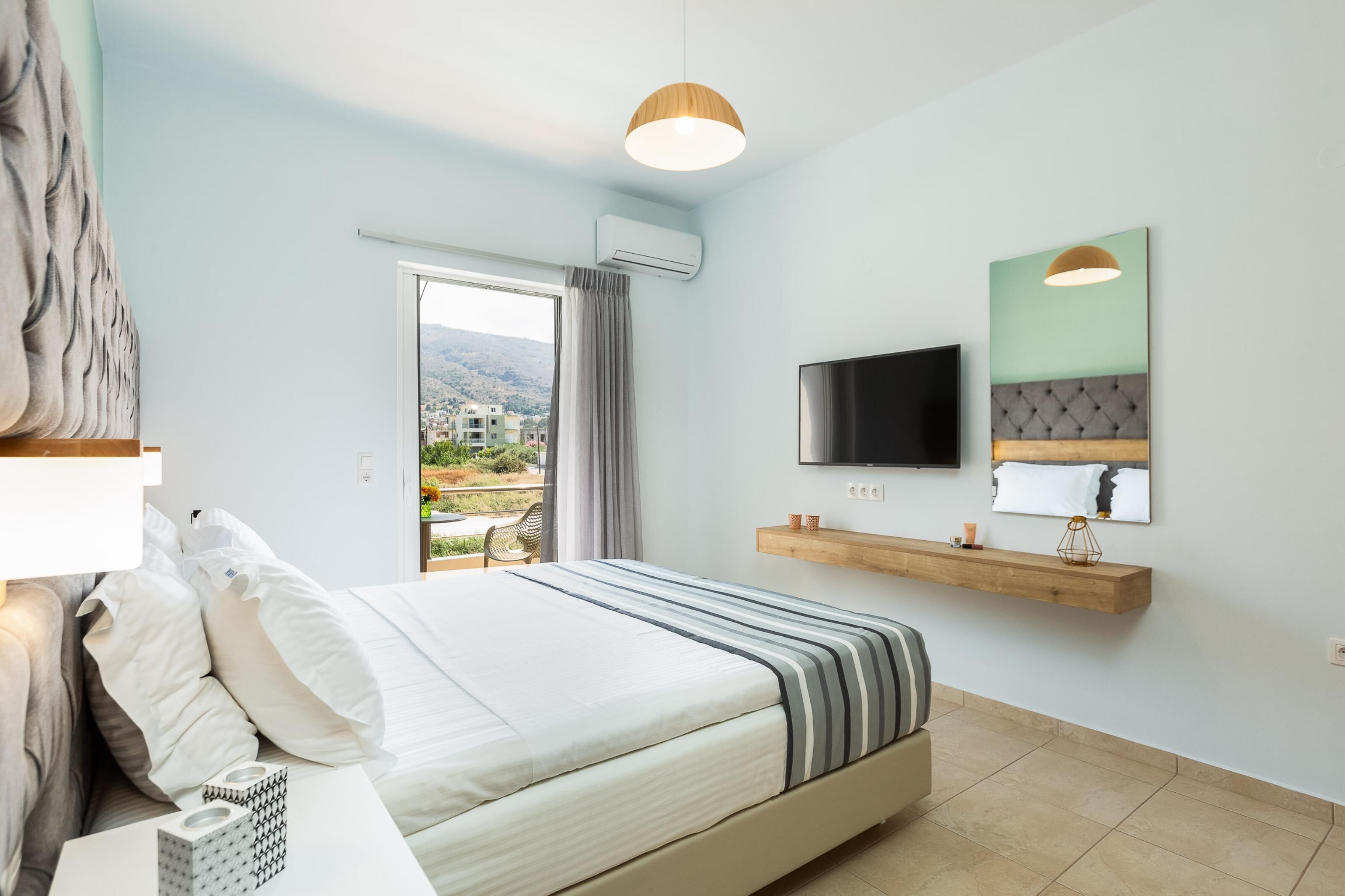 A modern hotel room with a large bed, a mounted TV, a wooden shelf with decorative items, and a view of a cityscape through a sliding door.