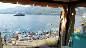 Blick auf die Küste mit einer großen Yacht in der Ferne, einem Strand mit Sonnenschirmen und Menschen, die das Wasser genießen.