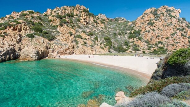 A coastal beach with clear turquoise water, rocky cliffs, and a sandy shore.