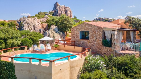 A pool with lounge chairs, a stone building with a red roof, and a rocky mountain backdrop.