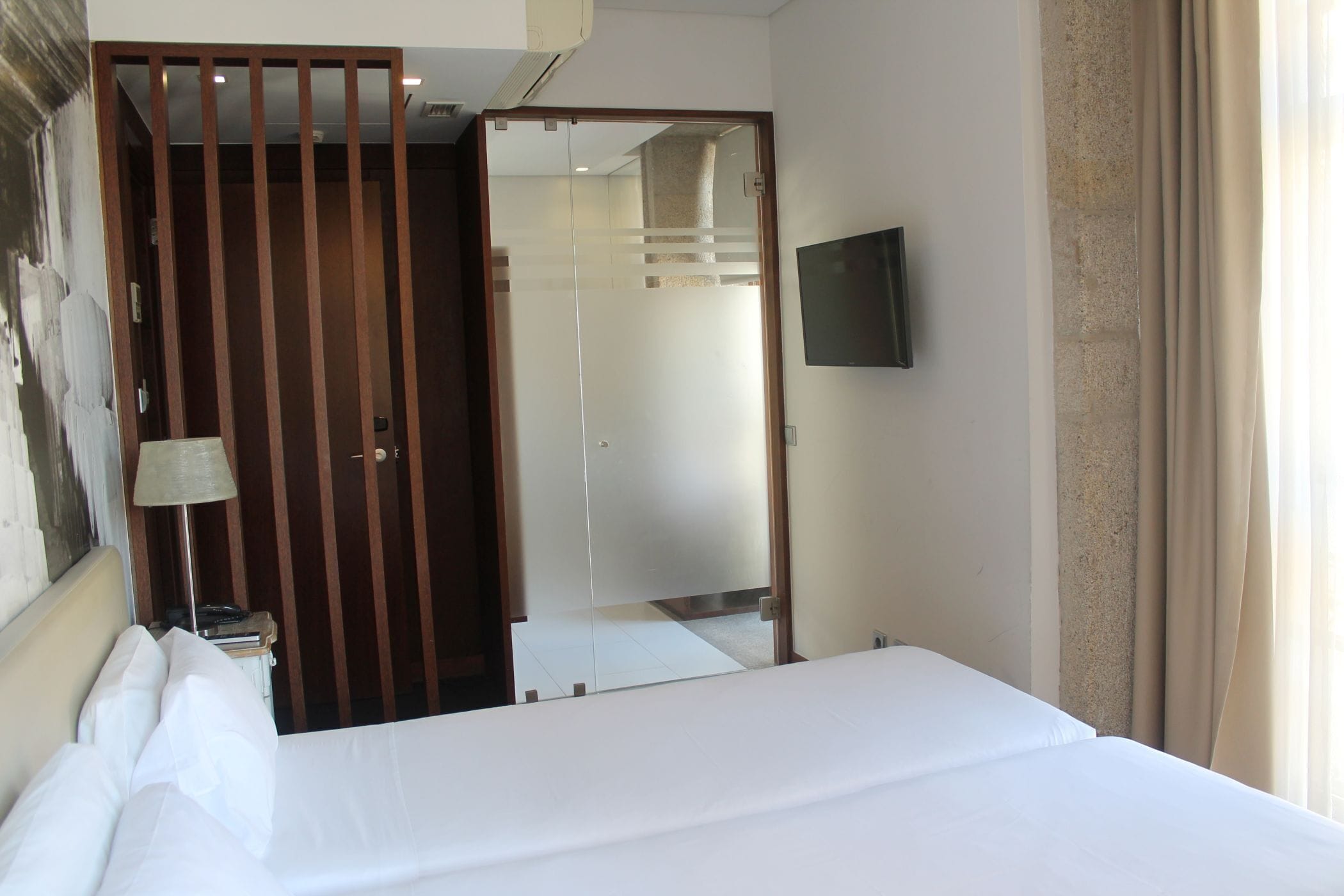 Habitación de hotel con cama, televisor montado en la pared, separador de madera y un espejo que refleja la habitación.