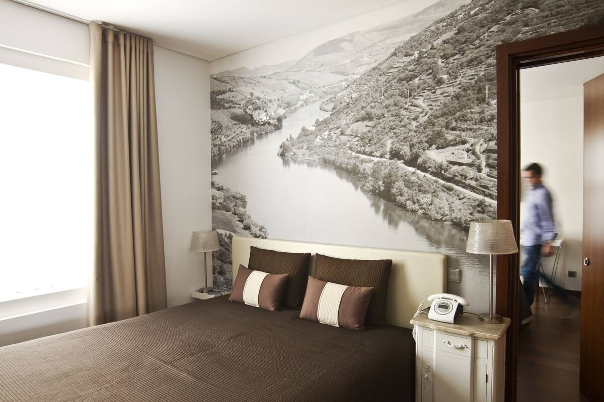 A hotel room with a large bed, two bedside lamps, a telephone, and a wall mural of a river.