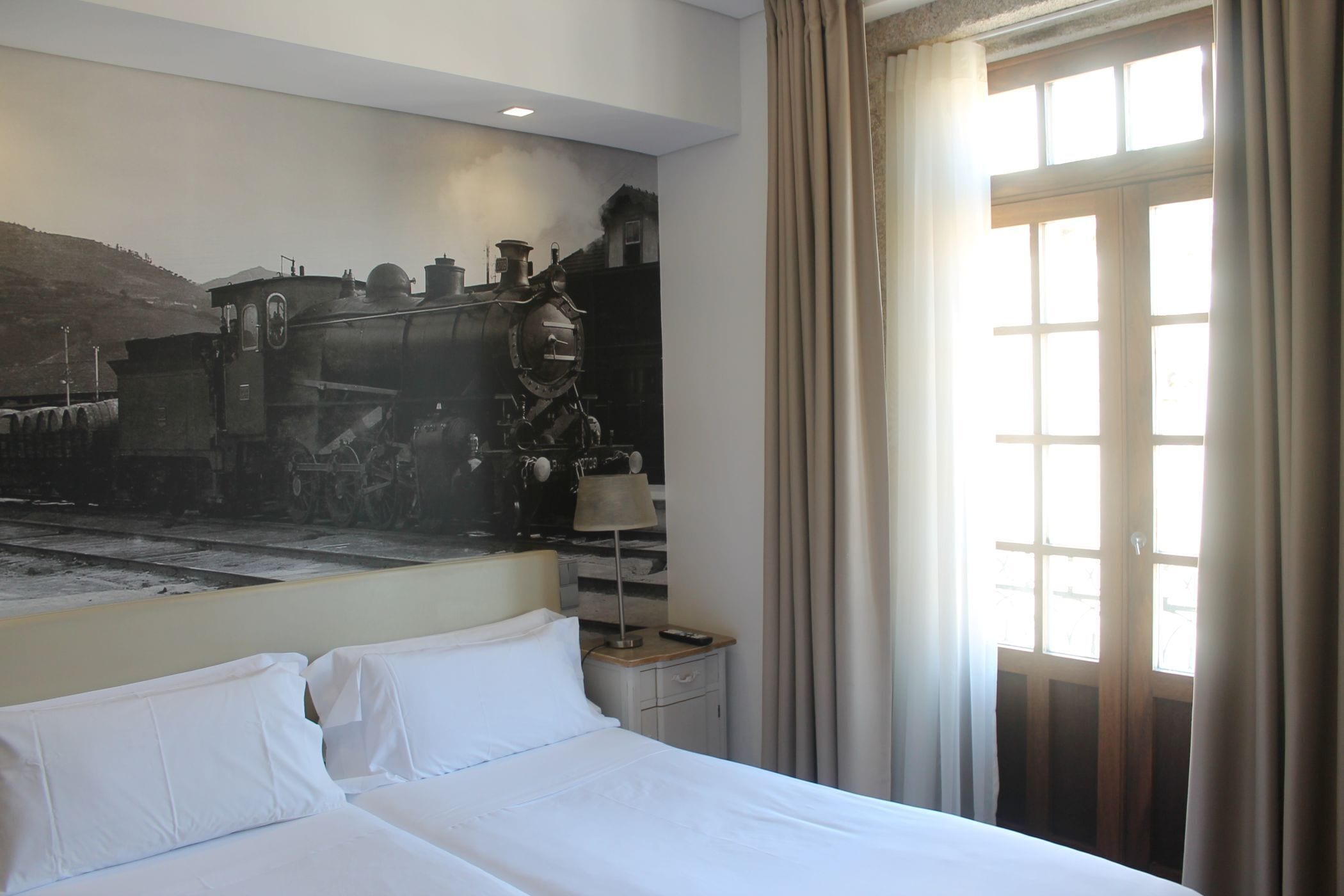 Quarto de hotel com uma cama grande, uma mesa de cabeceira com abajur e uma janela com cortinas.