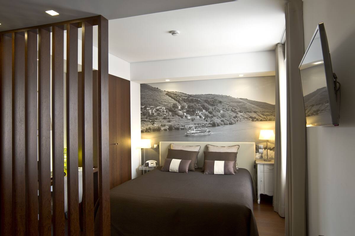 A hotel room with a bed, a nightstand, a lamp, a telephone, and a large landscape painting on the wall.