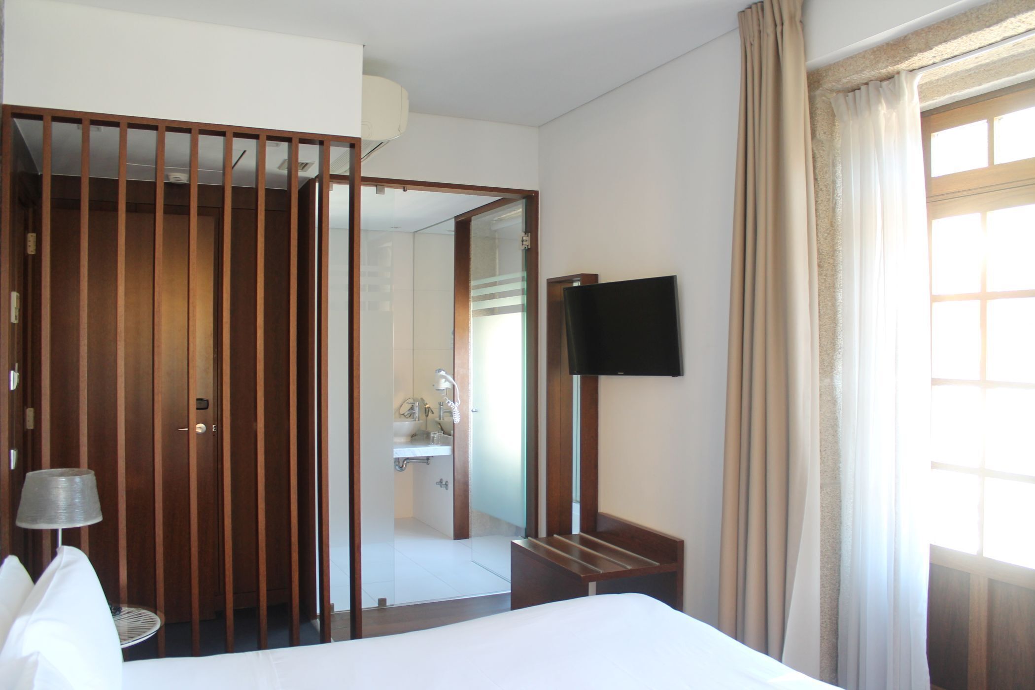 A hotel room with a bed, a television mounted on the wall, a wooden wardrobe, and a bathroom visible through an open door.