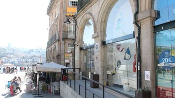 Un café al aire libre con mesas y sillas, un edificio de piedra con arcos y una vista de la ciudad al fondo.