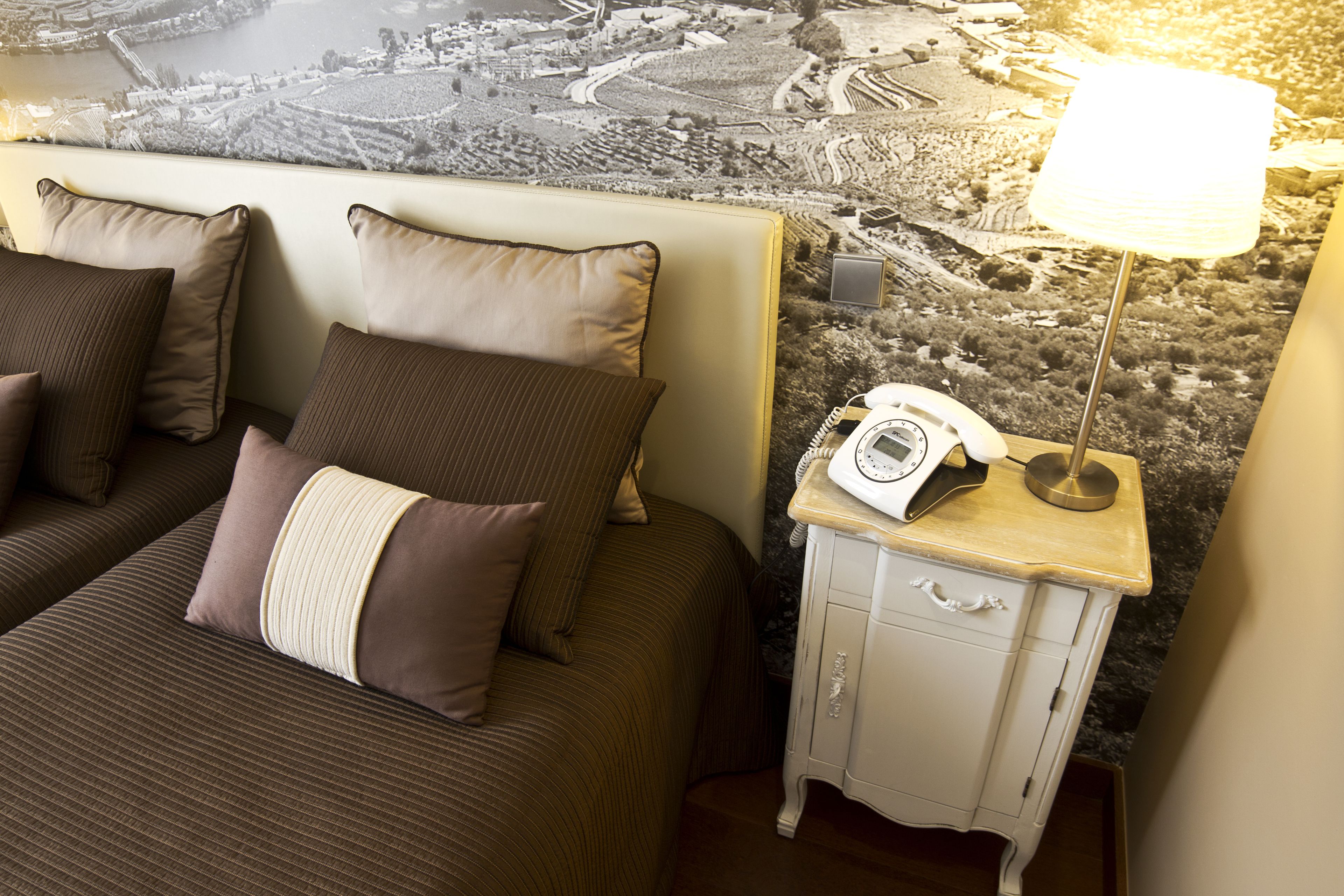 A hotel room with a bed, pillows, a nightstand with a lamp, and a wall mural.