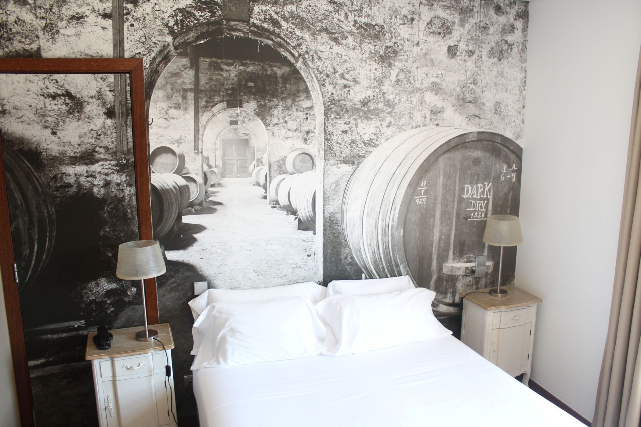 Une chambre d’hôtel avec un grand lit, une table de chevet, une lampe et une fresque murale représentant des tonneaux de vin.