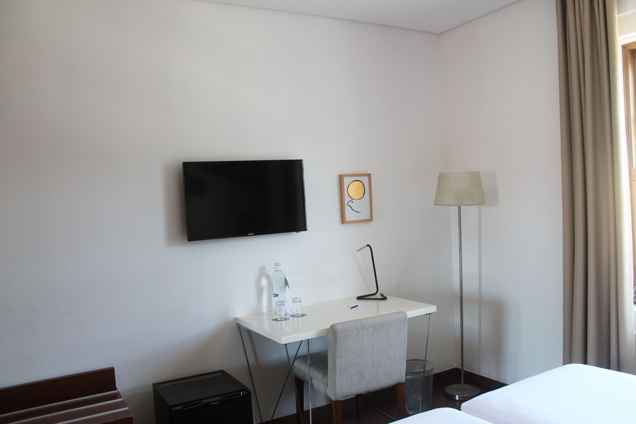 A hotel room with a bed, a desk with a chair, a TV, a lamp, and a small table with a bottle and glasses.