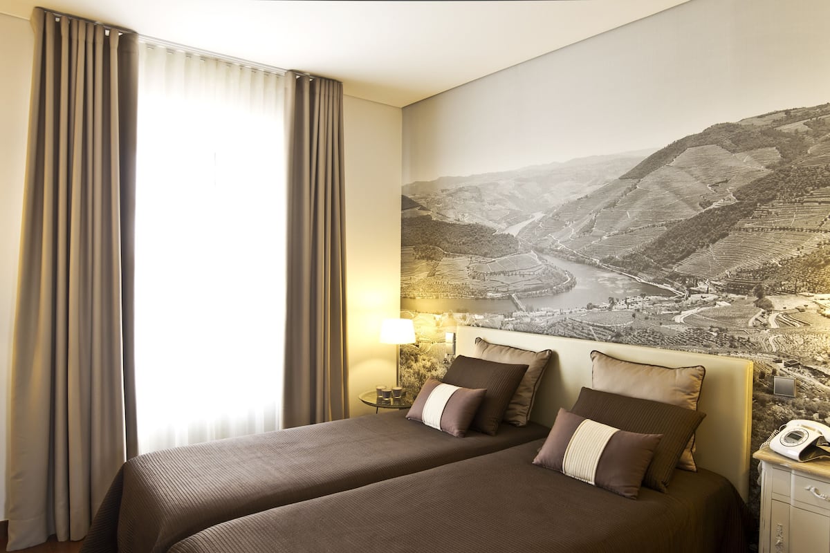 A hotel room with two beds, a large landscape mural on the wall, a bedside table with a lamp, and a window with curtains.