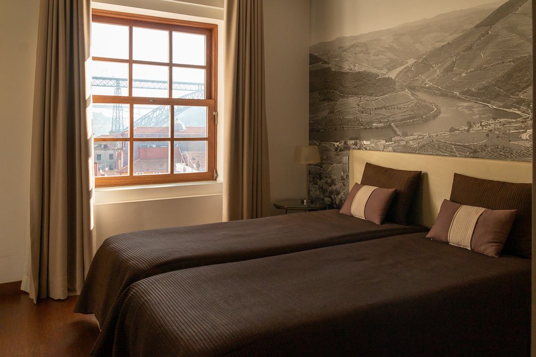 Quarto de hotel com duas camas, uma janela grande com cortinas, mesa de cabeceira com abajur e um mural com paisagem na parede.