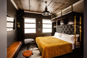 Habitación de hotel compacta con literas, un escritorio pequeño, una silla y un ventanal que deja entrar luz natural.