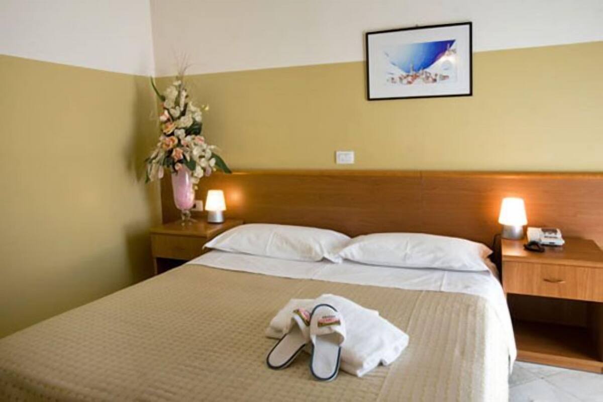 A hotel room with a bed, pillows, a nightstand with a lamp, a vase with flowers, and a framed picture on the wall.