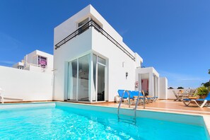 A modern building with a swimming pool, sun loungers, and a clear blue sky.