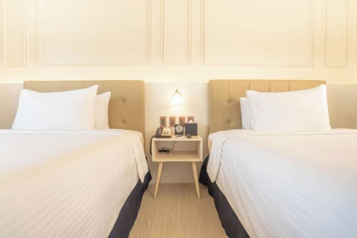 A hotel room with two beds, a bedside table with a lamp, and a wall-mounted light fixture.