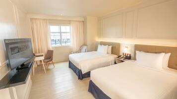 Habitación de hotel con dos camas, un escritorio con televisor, una silla, un ventanal con cortinas y una mesita de noche con lámpara.
