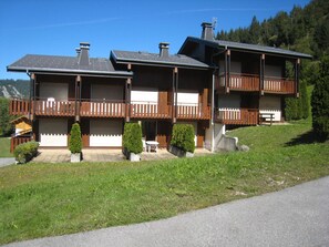 Un immeuble moderne à plusieurs étages, doté de balcons en bois, d’une entrée couverte et d’une allée bordée de pelouse.