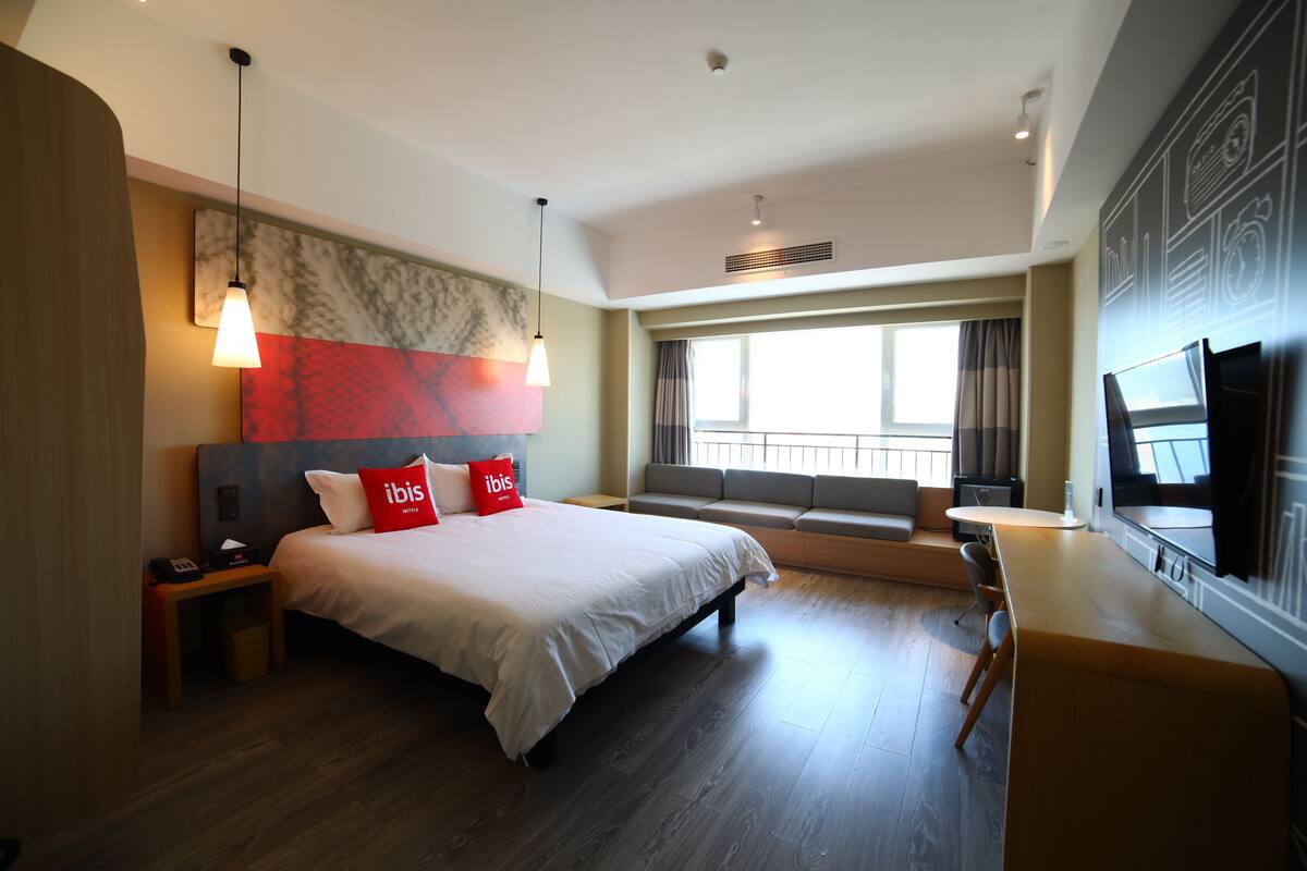 A modern hotel room with a large bed, a desk, a television, and a large window with curtains.
