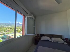 A bedroom with a bed, a nightstand, a lamp, and a window with a view of a landscape. - T2 Le frangipanier downtown, south of Reunion Island (saint louis)