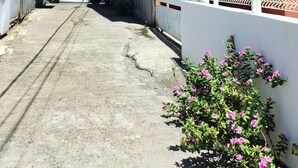 Une rue étroite, bordée de bâtiments des deux côtés, avec des unités de climatisation apparentes, une entrée fermée par un portail et une voiture garée sur le côté gauche.