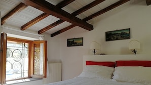 A bedroom with a wooden ceiling, a bed with white and red bedding, two wall-mounted lamps, and two framed pictures. - Baduerughe (Poggio Mirteto)