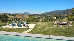 A pool area with lounge chairs and an umbrella, surrounded by a grassy lawn and a view of distant hills and houses. - Baduerughe (Poggio Mirteto)