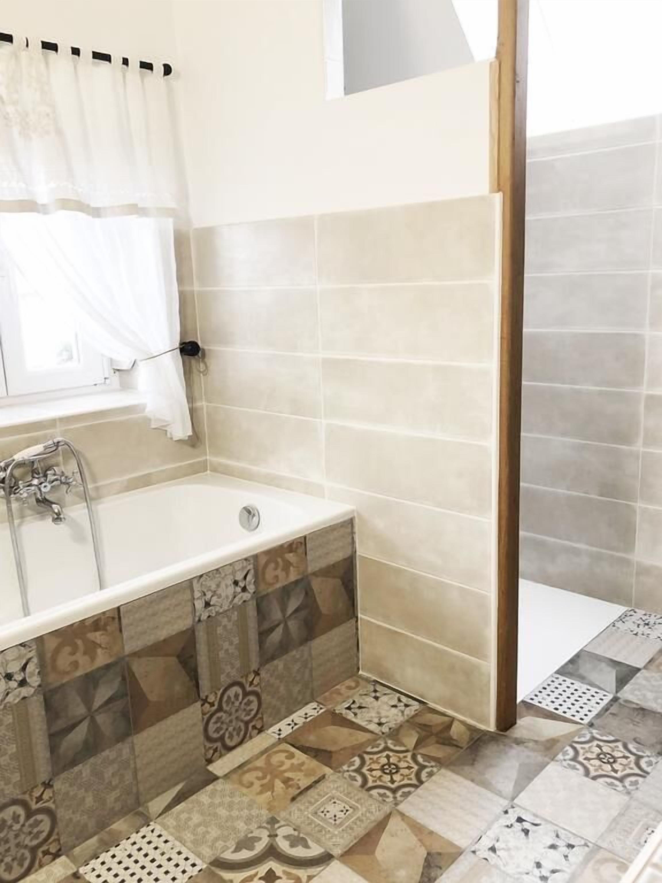 Ein Badezimmer mit einer Badewanne, einem Fenster mit weißem Vorhang und fugenwerkgefliesterten Böden und Wänden.