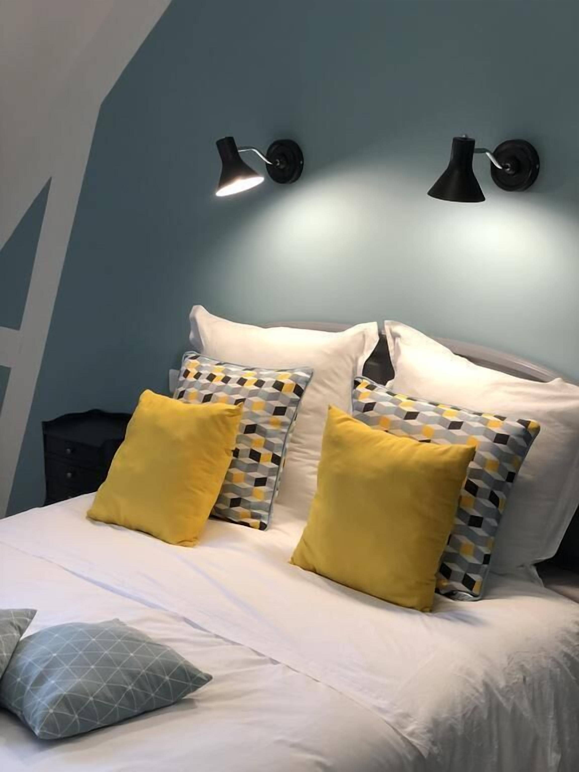 Una cama con ropa de cama blanca, dos almohadas amarillas y almohadas estampadas. Hay dos lámparas negras fijadas a la pared sobre la cama.