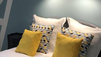 Una cama con ropa de cama blanca, dos almohadas amarillas y almohadas estampadas. Hay dos lámparas negras fijadas a la pared sobre la cama.