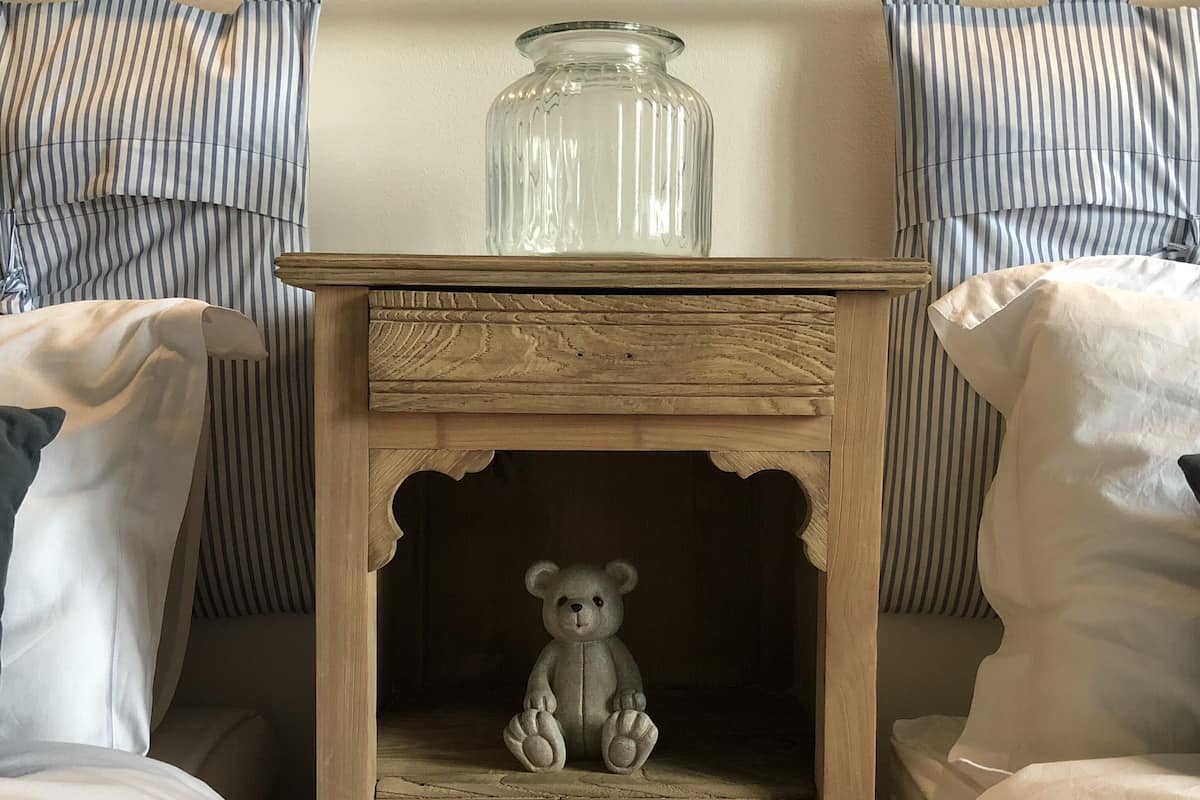 A wooden bedside table with a glass jar on top and a teddy bear inside.