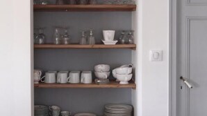 A kitchen with wooden shelves holding various kitchenware, a door to the right, and a light switch on the wall. - Il Etait Une Fois à L'Ouest (Plestan)