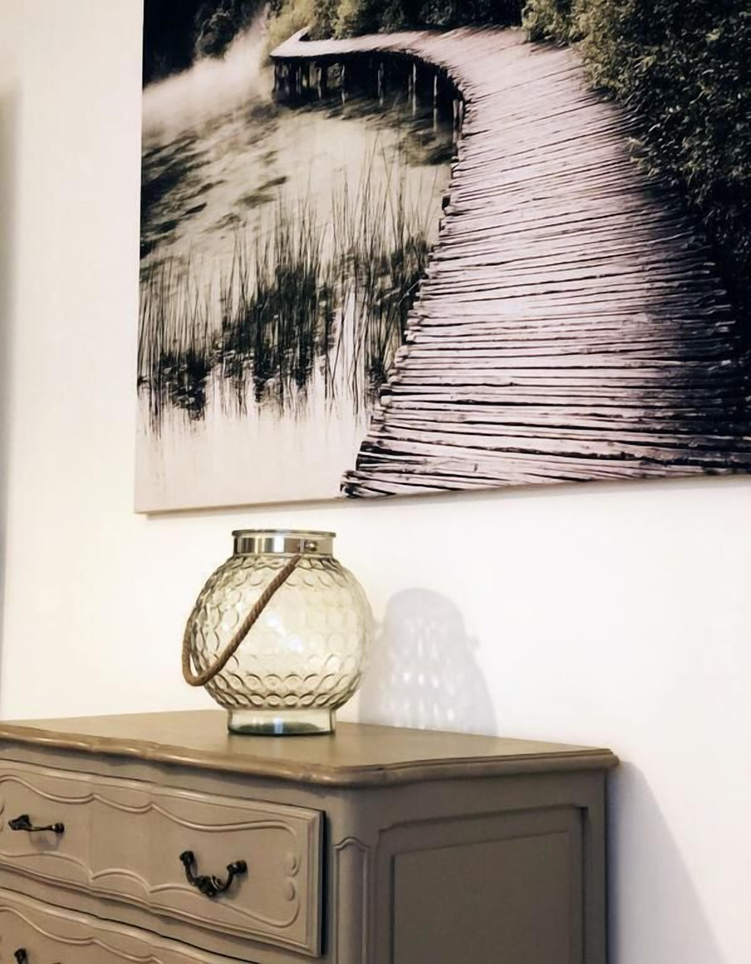 A wall with a painting of a wooden bridge over water, a decorative vase, and a dresser with drawers.