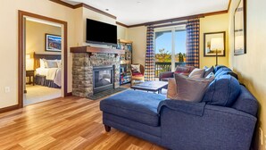 A living room with a stone fireplace, a flat-screen TV, a sofa, and a view of the outdoors. - 1st Fl Condo 3BR @ Stratton Sleeps 7 Pool Hot Tub (Stratton Mountain)