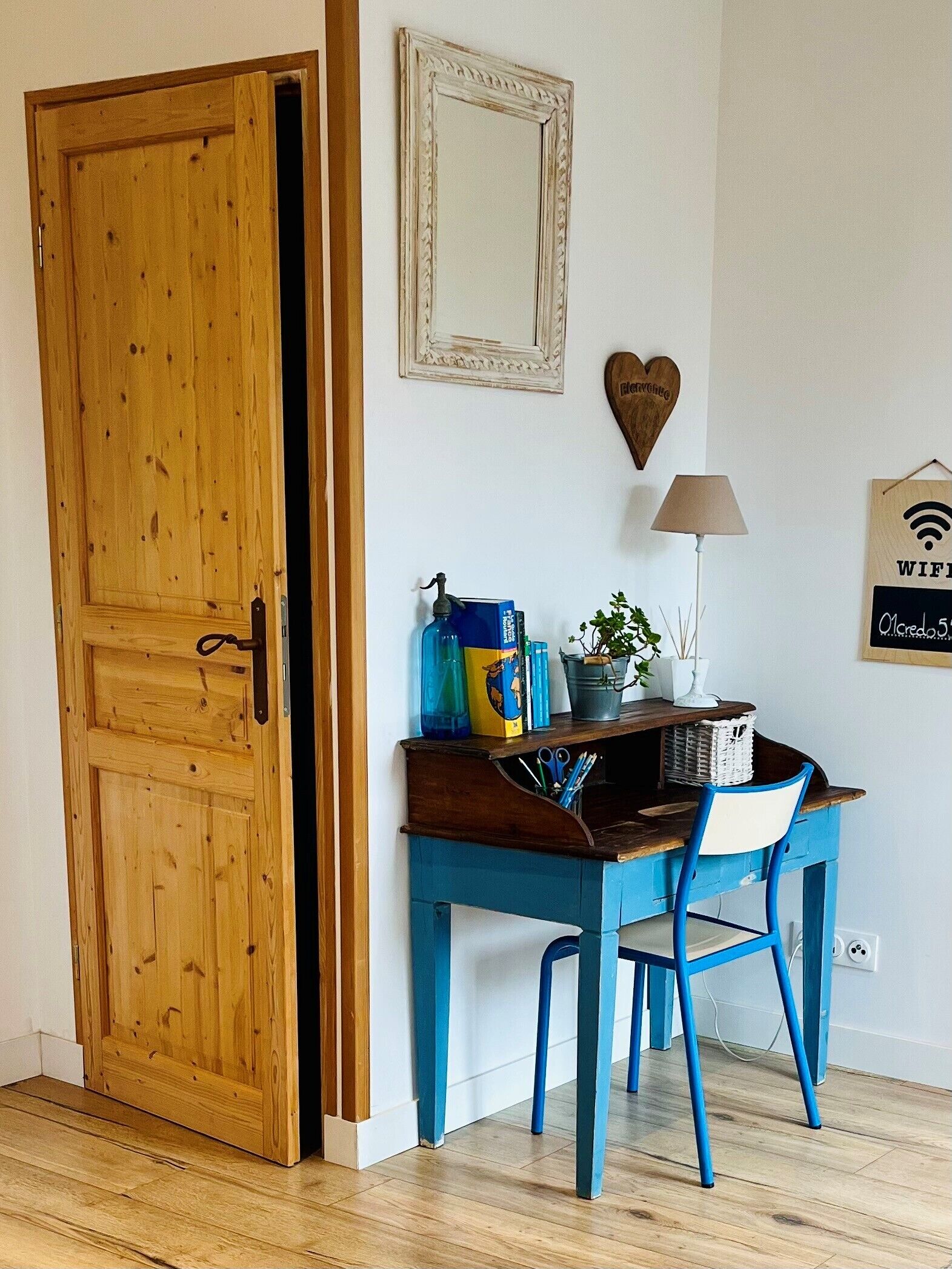 A room with a wooden door, a desk with a chair, a lamp, a heart-shaped decoration, and a wall-mounted sign indicating WiFi availability.