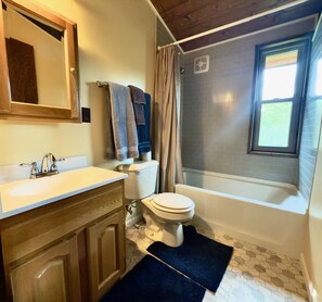 Un baño con tocador de madera, lavamanos blanco y un inodoro. Hay una cortina de ducha, una ventana y un perchero con toallas.