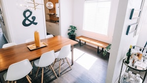 A modern dining area with a wooden table, white chairs, a bench, and a small kitchen in the background. - Designer Historic Duplex Minutes from Downtown (Phoenix)