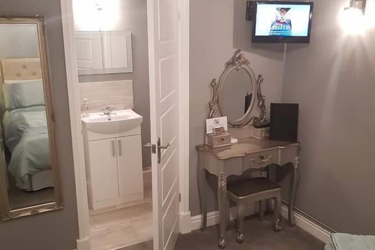 A bedroom with a bed, a desk with a mirror, a television, and a bathroom visible through an open door.