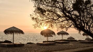 Una playa con sombrillas de paja y sillones de descanso, con vista despejada al atardecer sobre el agua.