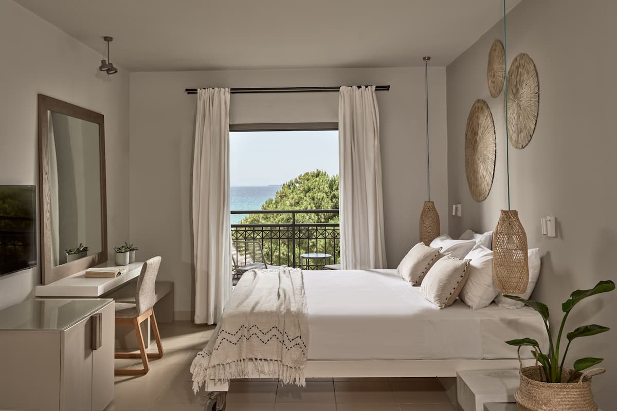 A bedroom with a bed, bedside table, and a desk with a chair. There is a balcony with a view of the sea.
