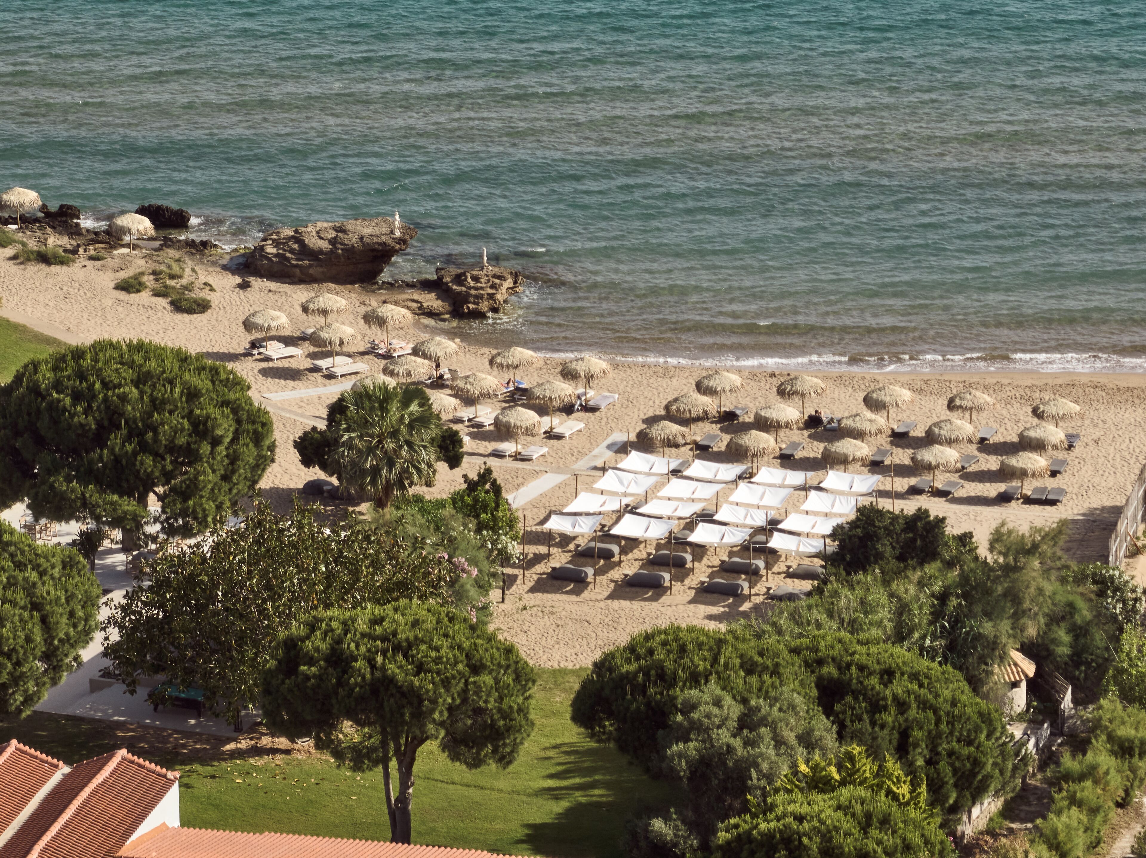 A beach with rows of sunbeds and thatched umbrellas, surrounded by greenery and rocky outcrops.