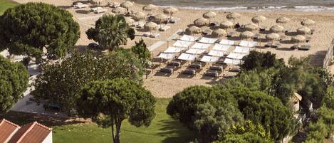 A beach with rows of sunbeds and thatched umbrellas, surrounded by greenery and rocky outcrops.