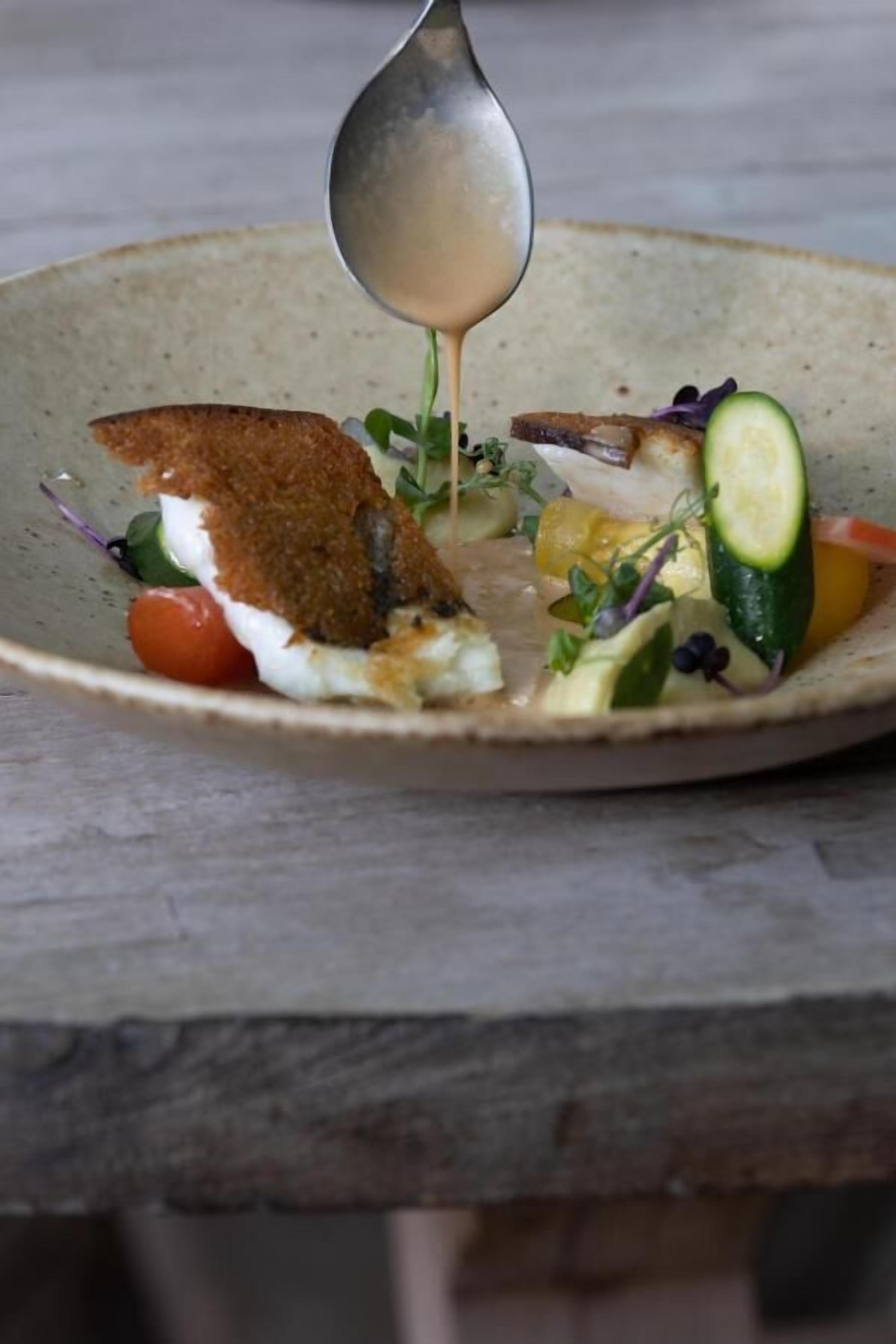 A plate with a variety of food items including a piece of breaded fish, vegetables, and a sauce being poured over the dish.