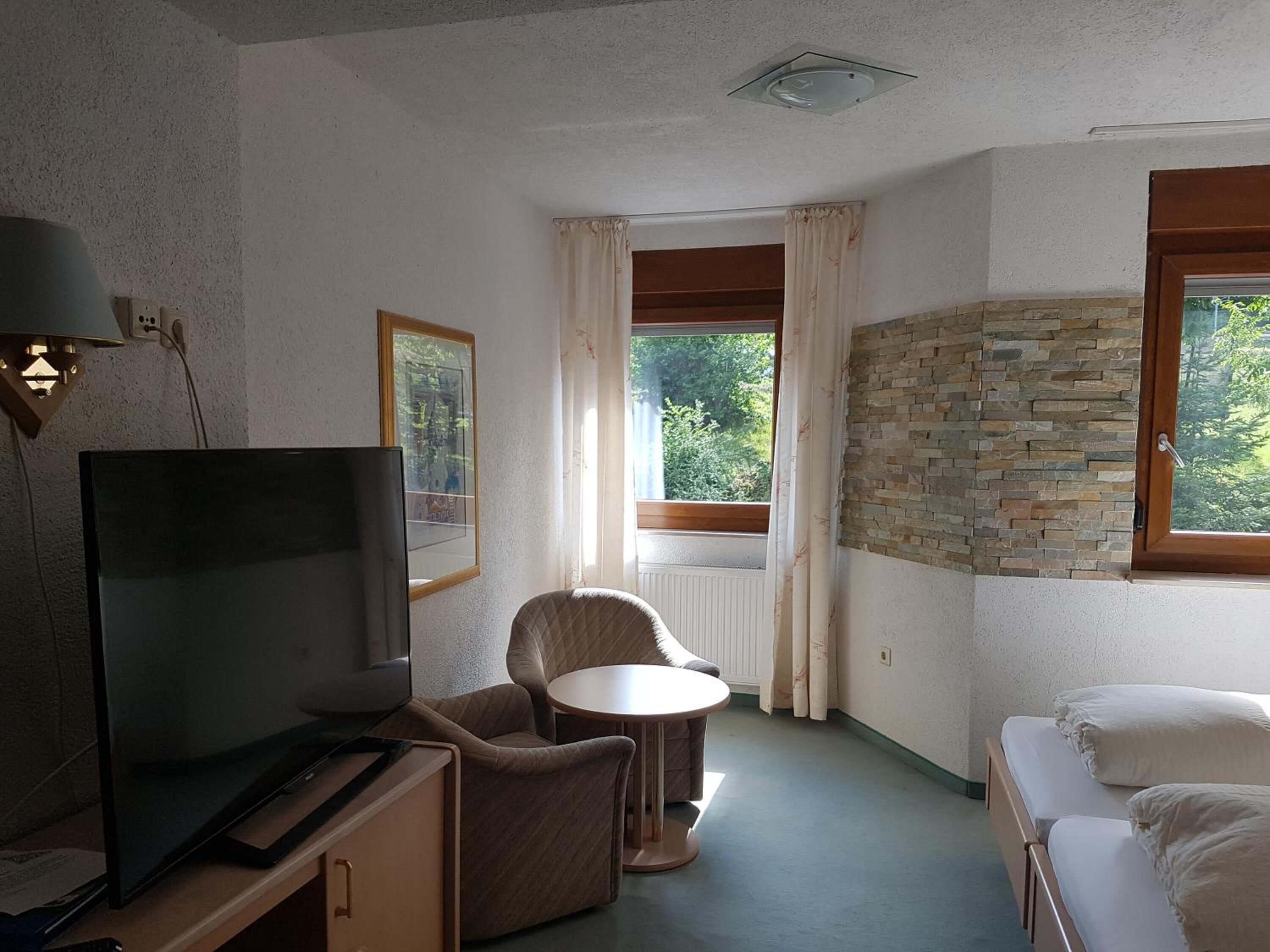 Une petite pièce agréable et confortable, avec un téléviseur, un fauteuil moelleux, une petite table ronde et un lit.