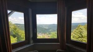 A room with a large window offering a view of a valley and hills. There are two chairs and a table with papers. - Hotel zur Engelsburg (Hausen)